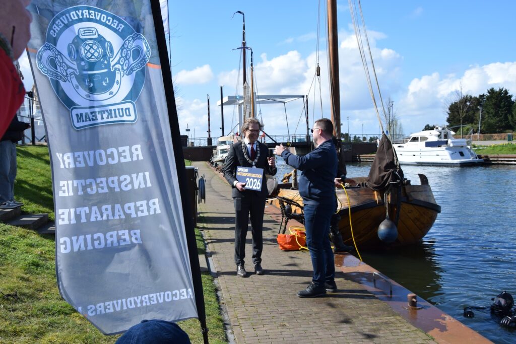 De loco-burgemeester met het opgedoken bord, met het duikteam in het water